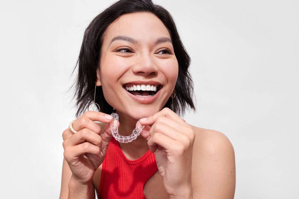 Woman smiling and holding a clear Candid Pro aligner in front of her teeth.